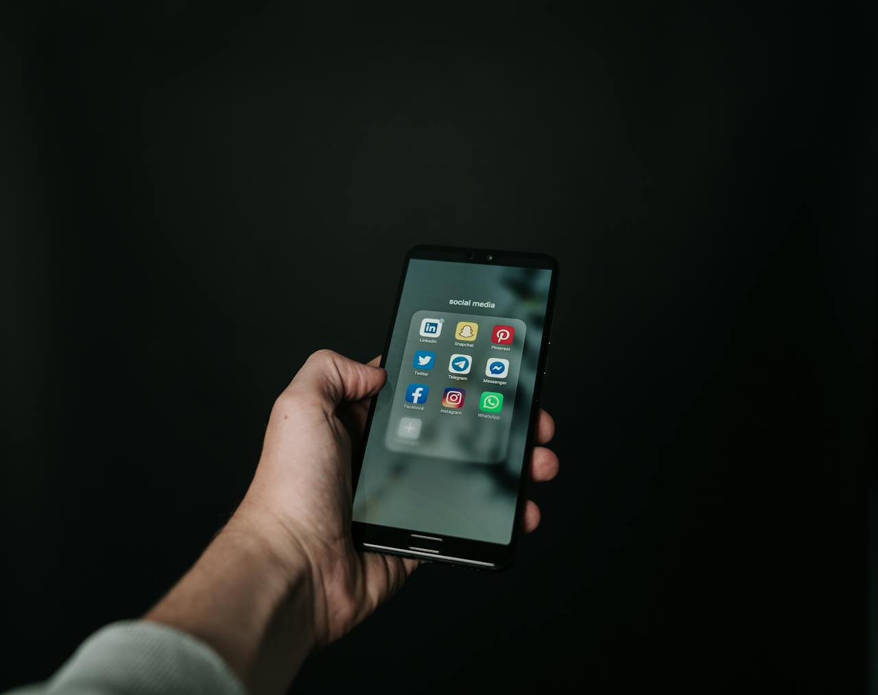 Hand holding a smartphone displaying a folder labeled 'Social Media' with app icons for LinkedIn, Snapchat, Pinterest, Twitter, Telegram, Messenger, Facebook, Instagram, and WhatsApp, against a dark background.