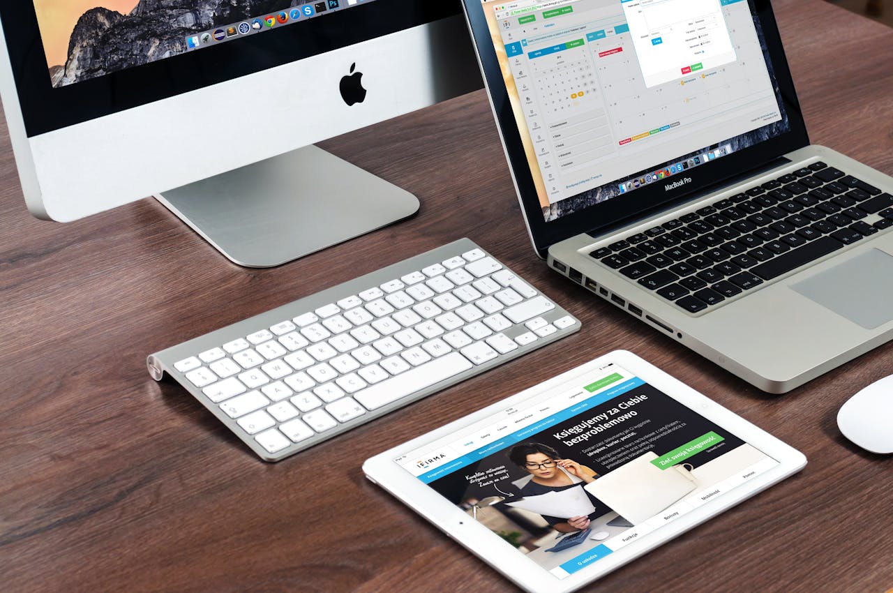 A sleek, organized workspace with an Apple iMac, MacBook Pro, wireless keyboard, and an iPad on a wooden desk, with a minimalist and modern aesthetic